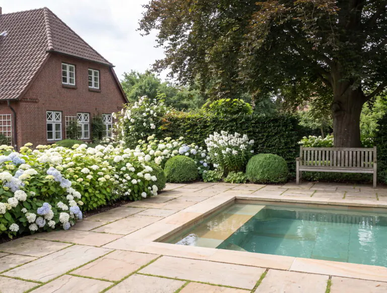 Pool mit Sandsteinrand in einem Landhausgarten mit weißen und blauen Hortensien, Buchsbaumkugeln, Holzbank und Klinkerhaus