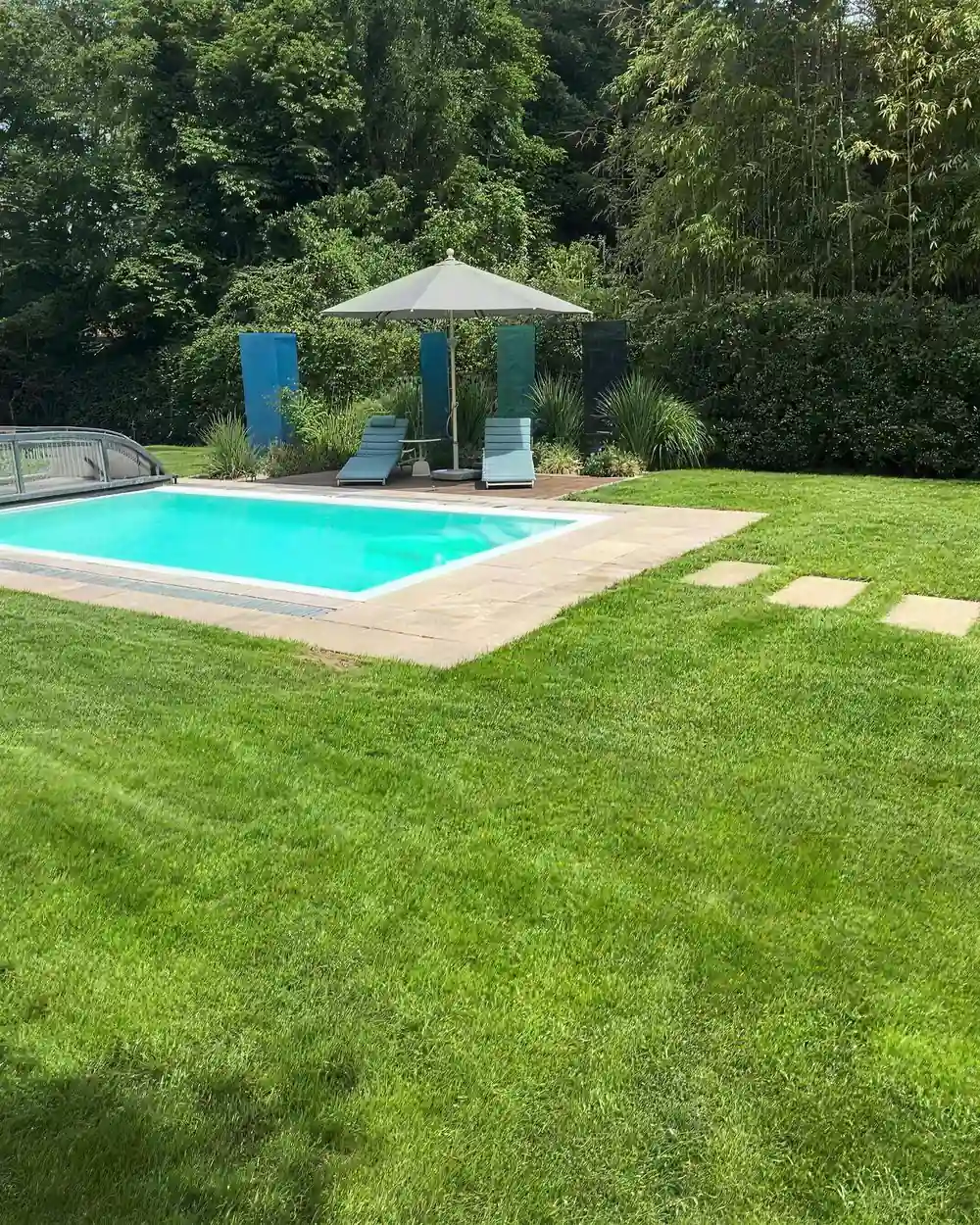 Rechteckiger Pool mit heller Umrandung in einem Rasengarten, daneben Sonnenliegen unter einem Sonnenschirm