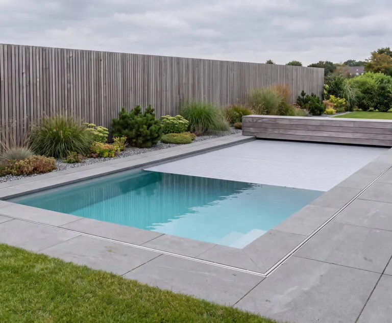 Pool mit halb eingefahrener Lamellenabdeckung, grauen Betonplatten, Sitzbank aus Holz und Sichtschutz aus Lärchenholz an einem bedeckten Tag