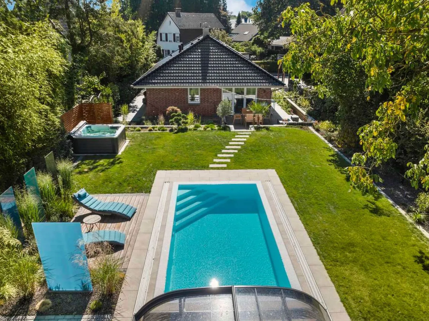 Luftaufnahme eines Gartens mit rechteckigem Pool, Whirlpool hinter einem Sichtschutz aus Holz, Holzdeck mit Liegen und Terrasse am Haus