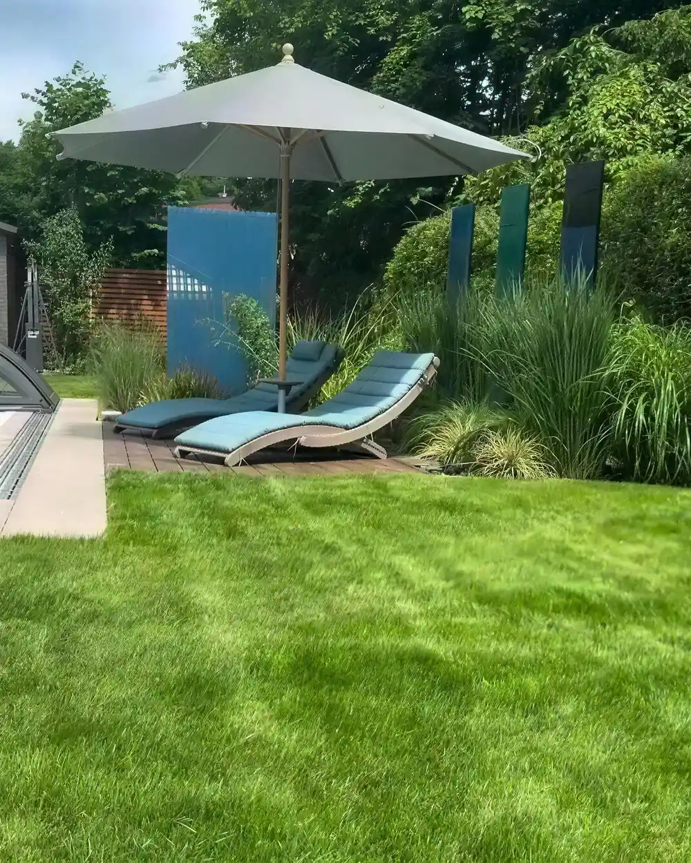 Sonnenliegen auf einem Deck am Poolrand, dahinter Gräser und ein Sonnenschirm